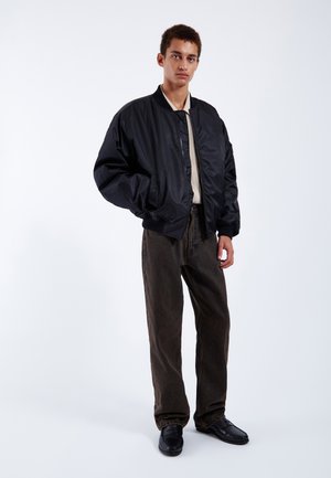 Black bomber jacket with a shiny finish, ribbed cuffs, beige shirt underneath, brown pants, black shoes, and a minimalist backdrop.