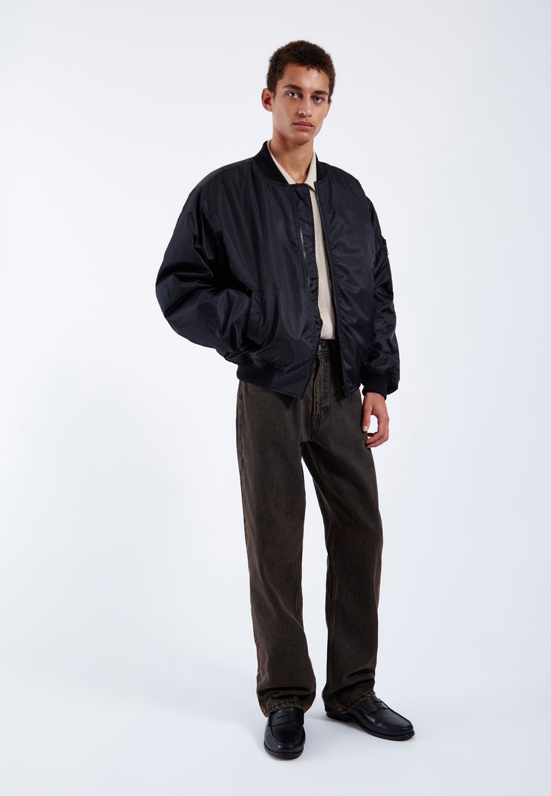 Black bomber jacket with a shiny finish, ribbed cuffs, beige shirt underneath, brown pants, black shoes, and a minimalist backdrop.
