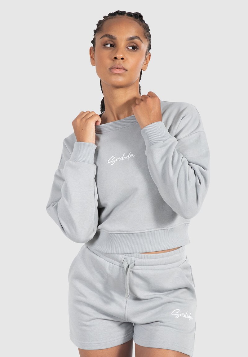 Lys grå cropped sweatshirt med ribstrikkede manchetter og talje, med "Smiledex" logo i hvid. Sammen med matchende grå shorts.