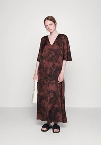 Vestido longo com padrão floral castanho, decote em V, mangas com metade do comprimento e textura suave. O modelo segura uma bolsa branca e usa sandálias pretas tipo slide.