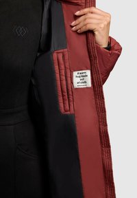 Padded bunda s vínovým exteriérom a čiernou podšívkou. Obsahuje prešívané panely a textilnú nálepku, na ktorej je napísané "Teplé objatie z látky."