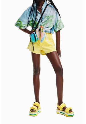 Osoba ve modré košili s tropickým vzorem, žlutých vysokých šortkách, na platformových sandálech a s brašnou přes rameno, s pletenými vlasy.