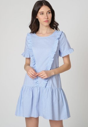 Mujer vistiendo un vestido de cuadros en azul claro con volantes en el frente, las mangas y el dobladillo, de pie con las manos entrelazadas.