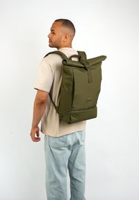 Zaino roll-top in tessuto verde oliva, con spallacci regolabili, tasche laterali e chiusura frontale. Forma rettangolare moderna e superficie liscia.