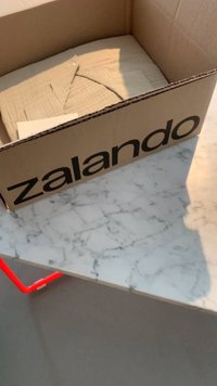 Scatola di cartone Zalando con logo nero contenente tessuto beige piegato, su una superficie in marmo con accentuazioni di supporto in metallo rosso.