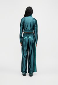 Γυναίκα όρθια, φοράει γυαλιστερό τζάκετ αθλητικό σε τυρκουάζ χρώμα και αντίστοιχο παντελόνι με πλατιά μπατζάκια, με λογότυπα Puma στις πίσω τσέπες και στις πλευρές.