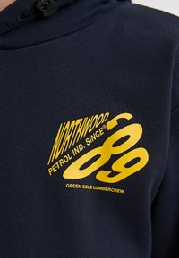 Námořnická modrá mikina s kapucí, která má velký žlutý grafický prvek s textem "NORTHWOOD PETROL IND. SINCE 89" a "GREEN GOLD LUMBERCREW." Měkká textilní struktura.