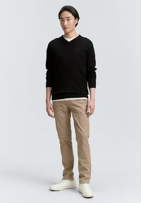 Maglione nero con scollo a V, pantaloni chino color cuoio e sneakers bianche indossati da un modello maschile in piedi davanti a uno sfondo neutro. Design semplice, senza motivi.