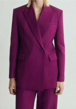 Femme portant un blazer magenta ajusté avec un pantalon assorti, sur un fond clair uni.