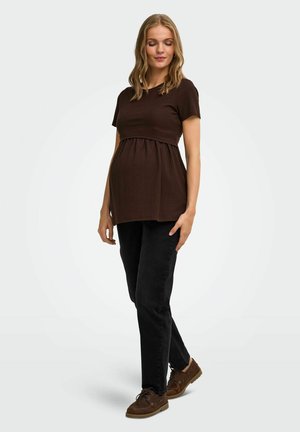 Femme enceinte portant un haut marron à manches courtes, un pantalon noir et des chaussures marron, debout les yeux fermés sur un fond clair.