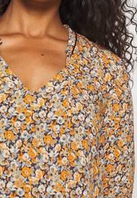 Blomstret bluse med V-hals, der har en blanding af orange, gule og blå blomster på en mørk baggrund. Letvægtsstof med tekstureret finish.