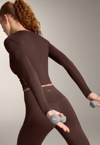 Vrouw in bruin sportkleding die 1 kg grijze halters optilt, gezien van achteren, tegen een neutrale beige achtergrond.