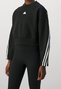 Felpa nera cropped realizzata in un materiale morbido. Presenta un logo bianco sul petto e tre strisce bianche lungo le maniche.