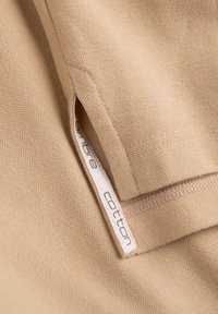 Béžová bavlněná tkanina s texturovaným povrchem. Na švu je bílá cedulka s nápisem "ombre cotton", která ukazuje na čisté zakončení.