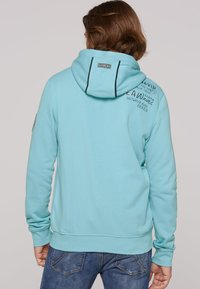 Light blue hoodie with a drawstring hood, ribbed cuffs, and waistband. Features text and patches on the back. Soft, textured fabric.