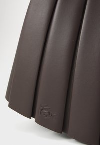 Section de jupes en cuir plissées marron foncé avec logo crocodile embossé près de l'ourlet sur un fond clair.