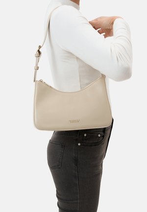 Beige leather shoulder bag with zipper and adjustable strap worn on right shoulder over white long-sleeve shirt and black jeans.