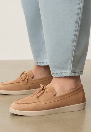 Jean bleu clair porté avec des mocassins marron clair à enfiler, ornés d'un nœud et de semelles blanches en caoutchouc sur un sol lisse.