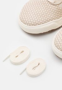 Beige színű sportcipők texturált szövet felsőrésszel, fehér gumitalppal, és két feltekeredett krémszínű fűzővel, amelyek a cipők alatt helyezkednek el.