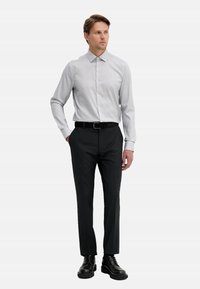 Camicia grigia chiaro con bottoni, maniche lunghe, abbinata a pantaloni neri e scarpe nere con lacci. Tessuto liscio con silhouette aderente.