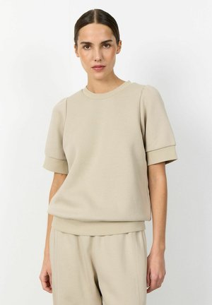 Žena s tmavými vlasmi v béžovej krátkosleeve mikine a zodpovedajúcich nohaviciach, stojaca pred jednoduchým bielym pozadím.