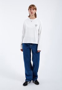 Long-sleeve white shirt with a small black heart logo on the left chest, paired with loose blue jeans and black sneakers.