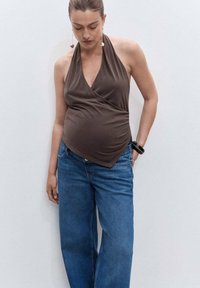 Žena oblečená v hnedom bezrámovom halterneku a vo voľných modrých džínsoch, stojí na pozadí bielej steny a pozerá dole.