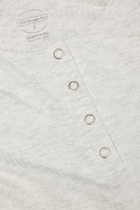 Camisa de tela acanalada gris claro con cuatro botones de presión de metal plateado y etiqueta impresa que muestra la marca, la talla y el origen.