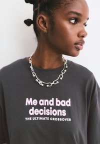 Jonge vrouw met gevlochten haar in een donker shirt met de tekst "Ik en slechte beslissingen, de ultieme crossover" en een zilveren kralenketting.