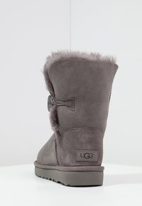 Stivale grigio in suede con fodera in pelliccia sintetica, caratterizzato da un bottone decorativo e una suola piatta in gomma. Etichetta del marchio visibile sul tallone.