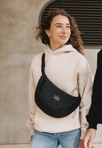 Bolso cruzado negro hecho de tela duradera, con un diseño semicírculo, correa ajustable y un pequeño parche con logo en la parte frontal.