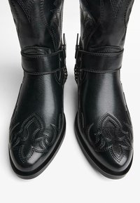 Črne usnjene kavbojske čevlje z zapletenimi šivi, kovinskimi sponkami in okroglo oblikovano konico. Gladka tekstura in nizka peta.