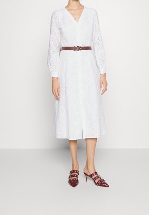 Femme portant une robe midi blanche à manches longues avec motif à œillets, ceinture marron et mules bordeaux à talons cloutés sur un fond uni.