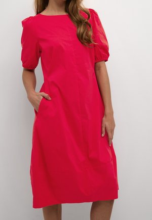 Kvinde iført en lys pink knælængde kjole med puffede korte ærmer, den ene hånd i lommen, stående mod en ensartet hvid baggrund.