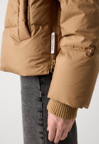 Gros plan sur une main dans une manche en tricot beige sous une veste rembourrée beige avec l'étiquette Marc O'Polo, portée sur un jean en denim gris.