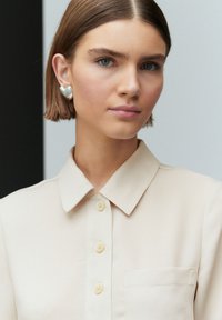 Béžová košile s špičatým límcem a předními knoflíky. Modelka má na sobě stříbrné náušnice ve tvaru srdce. Hladká textura, minimalistický design.