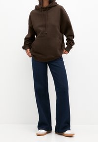 Felpa in cotone marrone con tasca frontale e cordini, abbinata a jeans svasati blu scuro e scarpe sportive bianche.