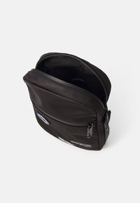 Černá látková kabelka na crossbody s uzávěrem na zip, která má přední nášivky, hladkou texturu a strukturovaný obdélníkový tvar.