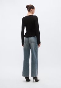 Bluza neagră cu mâneci lungi și margine din dantelă, asortată cu blugi evazați albastru deschis și cizme negre cu toc înalt. Modelul este cu fața spre spate, fundal minimalist.