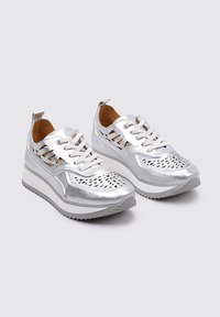 Zapatillas metálicas plateadas con un diseño de malla, perforaciones con patrón, cordones blancos y una suela blanca gruesa. Presenta una tira en la parte trasera para ayudar a calzarlas.