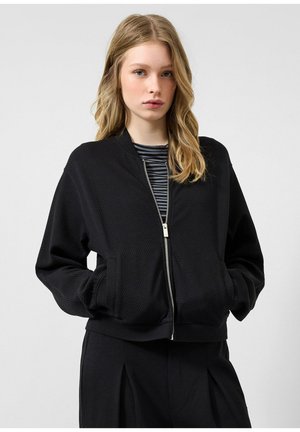 Junge Frau mit blondem Haar, die eine schwarze, texturierte Jacke mit Reißverschluss und schwarze Hosen über einem gestreiften Hemd trägt, steht vor einem einfarbigen Hintergrund.