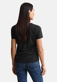 Γυναίκα με μακριά σκούρα μαλλιά που φοράει μαύρη κοντομάνικη μπλούζα και μπλε τζιν, γυρισμένη πλάτη προς ένα απλό φωτεινό φόντο.