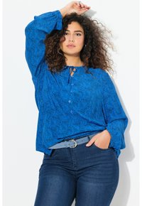 Blaue Bluse mit einem wirbelnden Muster, Knopfdetail und Krawattenhals. Getragen mit dunklen Jeans und einem hellblauen Gürtel. Lange Puffärmel.