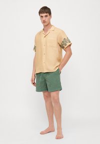 Camisa corta de manga corta color amarillo claro con detalles bordados verdes en las mangas, combinada con pantalones cortos de cuadros verdes. Ambos artículos están hechos de tela.
