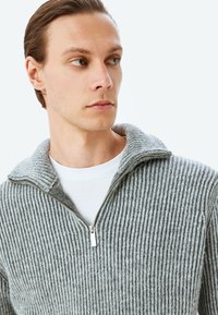 Grau gestreifter Reißverschluss-Pullover mit hohem Kragen, strukturiertem Strickstoff, ausgestattet mit einem silbernen Reißverschluss und über einem weißen Hemd getragen.