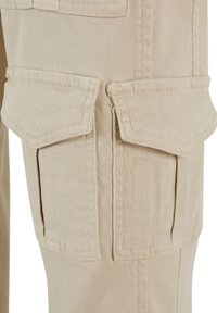 Beige cargo nadrág, amely két külső fedeles zsebbel rendelkezik a combokon, tartós anyagból készült, látható varrással és strukturált felülettel.