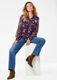Blusa trasparente viola con motivo floreale, maniche lunghe e scollo con fiocco, abbinata a jeans blu e stivali alla caviglia in suede marrone.