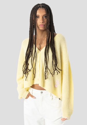 Frau mit langem geflochtenem Haar, die einen lockeren gelben Knopfpullover und weiße Hose trägt, steht vor einem schlichten hellen Hintergrund.