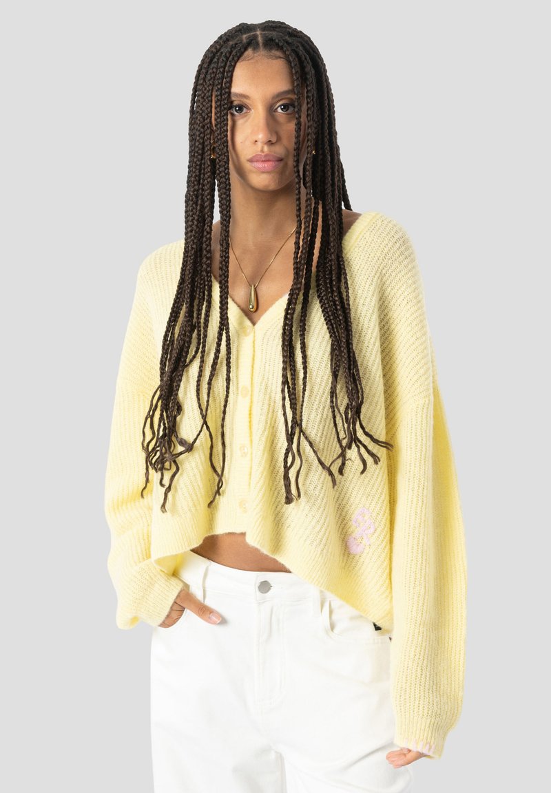 Femme aux longs cheveux tressés portant un pull jaune ample boutonné et un pantalon blanc, debout devant un fond clair uni.