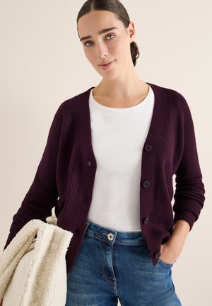 Burgunderfarbener gerippten Cardigan mit Rundhalsausschnitt, fünf Knöpfen und langen Ärmeln, kombiniert mit einem weißen Hemd und blauen Jeans. Hält eine cremefarbene strukturierte Tasche.